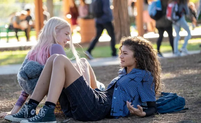 Two young women sit on the ground in a park, talking and laughing, with a blurred crowd in the background.