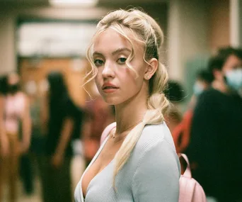 Character from Euphoria Season 2 with styled hair, wearing a light top, in a school hallway setting.