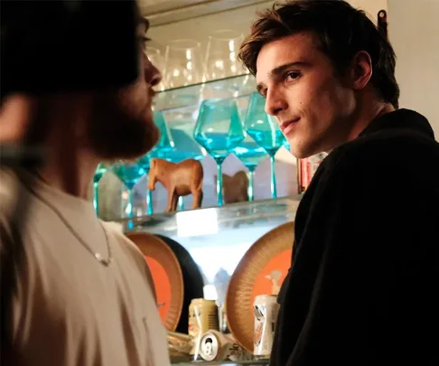 Two men in a bar-like setting with blue glasses on shelves; one smiles while the other faces away.