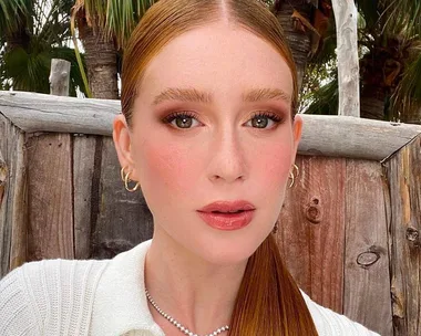 Woman with red hair and natural makeup, wearing a white top and gold earrings, posed outdoors against a wooden background.