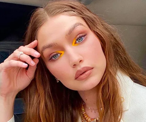 Person with sunkissed blonde hair, vibrant eye makeup, wearing neutral lipstick and a beaded necklace, posing thoughtfully.