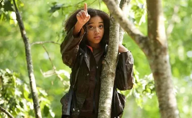 A character in The Hunger Games, wearing brown jacket, points upwards while standing by a tree in a forest.