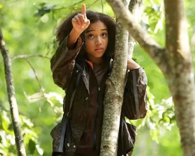 A character in The Hunger Games, wearing brown jacket, points upwards while standing by a tree in a forest.