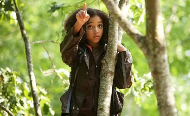 A character in The Hunger Games, wearing brown jacket, points upwards while standing by a tree in a forest.