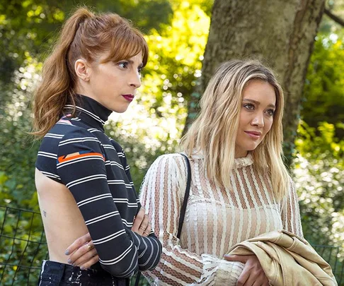 Two women stand outdoors in a shaded area, both wearing patterned tops, in a thoughtful pose.