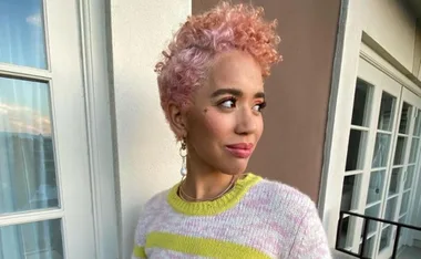 Person with short pink curly hair, wearing a striped sweater, looks to the side while standing on a balcony.