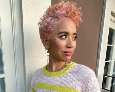 Person with short pink curly hair, wearing a striped sweater, looks to the side while standing on a balcony.