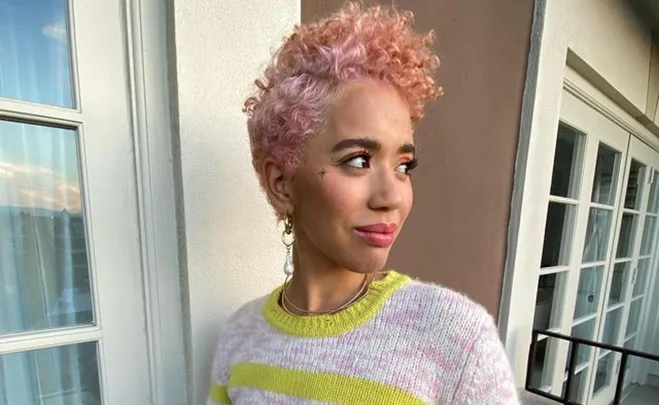 Person with short pink curly hair, wearing a striped sweater, looks to the side while standing on a balcony.