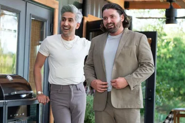 Two men smiling and standing outside a home; one wears a white shirt and the other a beige suit.