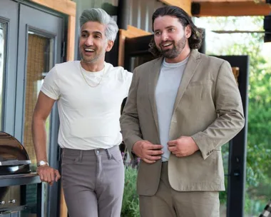 Two men smiling and standing outside a home; one wears a white shirt and the other a beige suit.