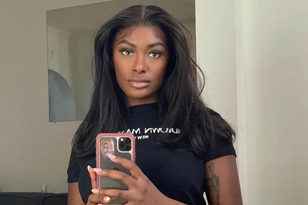 Woman taking a mirror selfie with a phone, wearing a black t-shirt, in a room with neutral walls.