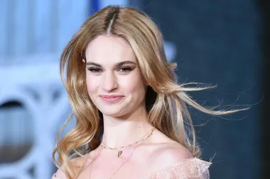 Lily James smiling with long blonde hair, wearing a light pink off-shoulder dress and necklace.