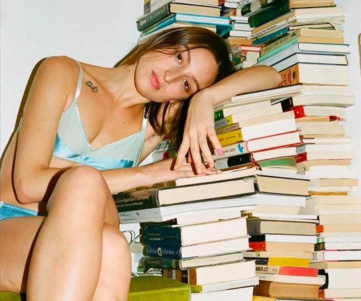 A woman in light blue lingerie leans against a stack of books, looking contemplative.