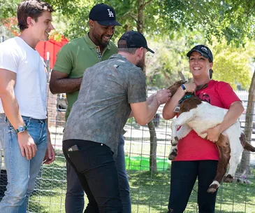 Four people smiling, one holding a goat, in a sunny outdoor setting near a fence.