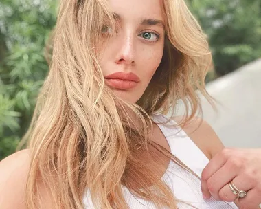 A woman with long blonde hair and freckles, wearing a white top, posing outdoors with greenery in the background.