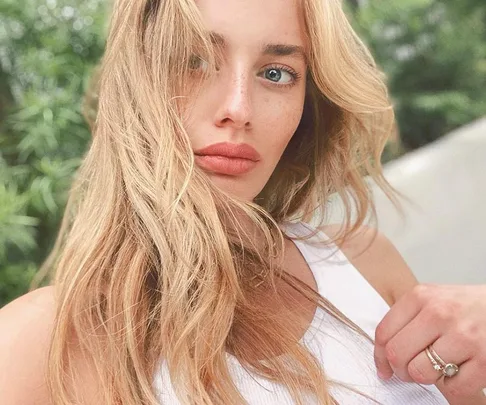 A woman with long blonde hair and freckles, wearing a white top, posing outdoors with greenery in the background.