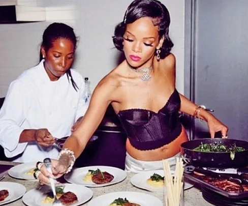 Two women plating food in a kitchen; one in a white coat, the other in a corset top.