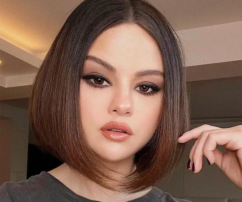 Selena Gomez with a sleek bob hairstyle, dramatic eye makeup, and nude lip color, posing indoors.