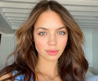 A woman with wavy brown hair and light makeup, wearing hoop earrings and a necklace, stands in a room with wooden beams.