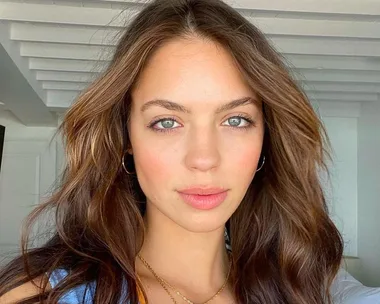 A woman with wavy brown hair and light makeup, wearing hoop earrings and a necklace, stands in a room with wooden beams.