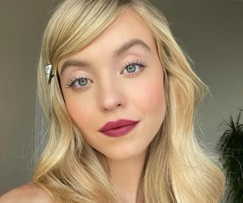 A person with long blonde hair and burgundy lipstick, featuring a makeup look with a hair clip.