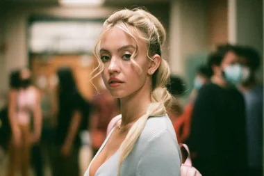 A character from "Euphoria" with blonde hair and a light blue top stands in a hallway, looking seriously at the camera.