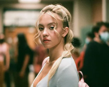 A character from "Euphoria" with blonde hair and a light blue top stands in a hallway, looking seriously at the camera.