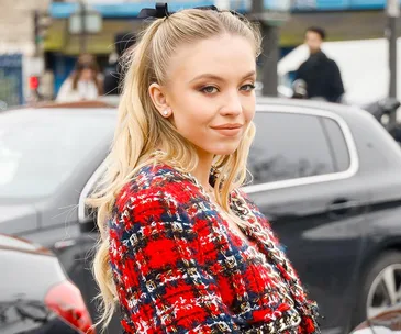 Young woman in checkered red and blue outfit, smiling, with long blonde hair, standing in front of parked cars.