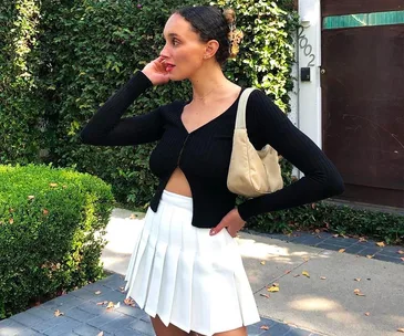 A woman in a black top and white skirt poses outdoors, holding a beige bag.