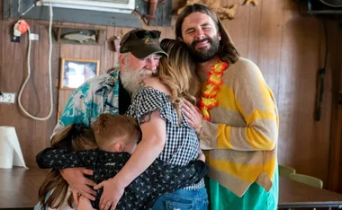 A bearded man and a person in a Hawaiian shirt hug two adults and a child in a warm, emotional group embrace.