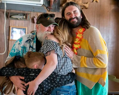 A bearded man and a person in a Hawaiian shirt hug two adults and a child in a warm, emotional group embrace.