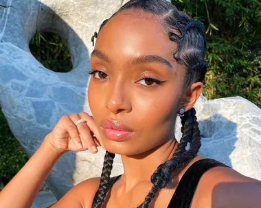 Person with braided hair and winged eyeliner poses outdoors, touching their chin, with a serene expression.