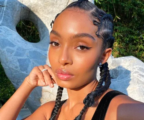 Person with braided hair and winged eyeliner poses outdoors, touching their chin, with a serene expression.