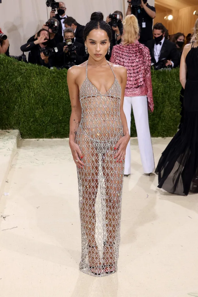 A person in a sheer, diamond-patterned dress poses on a beige carpet with photographers in the background.