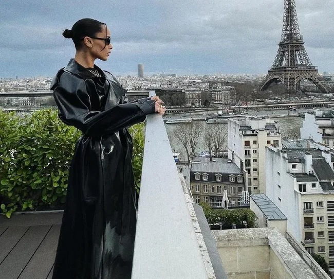 Person in black coat and sunglasses stands on a terrace overlooking the Eiffel Tower in Paris.