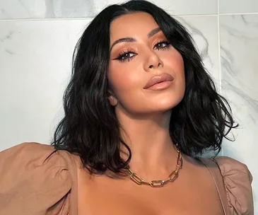 Person with dark hair wearing orange eyeshadow, a gold chain necklace, and a beige top, posed in front of a marble wall.