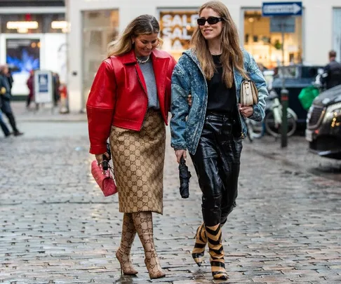 Two stylish women walking on a cobblestone street, one in a red jacket and Gucci print, the other in denim and leather.