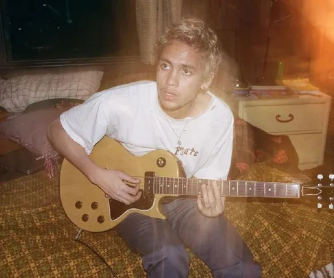 Person with short hair playing an electric guitar on a bed inside a cozy, dimly-lit room.