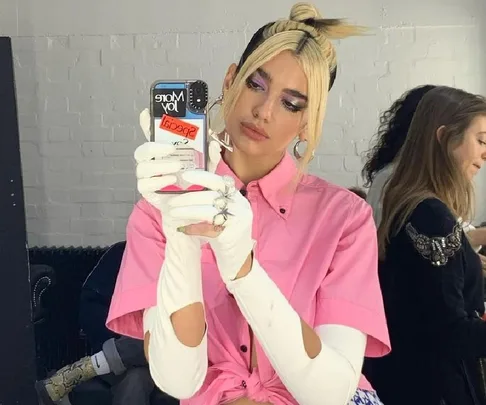 Singer in pink shirt and white gloves takes a mirror selfie with a phone in a colorful case.