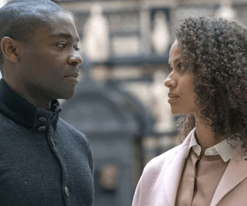 Two people, one in a coat and the other in a pink blazer, stand facing each other, set against a blurred background.