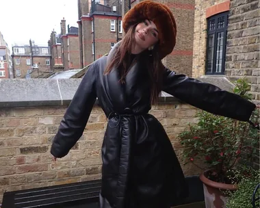 Woman in a black leather coat and brown fur bucket hat poses joyfully on a brick terrace in an urban setting.