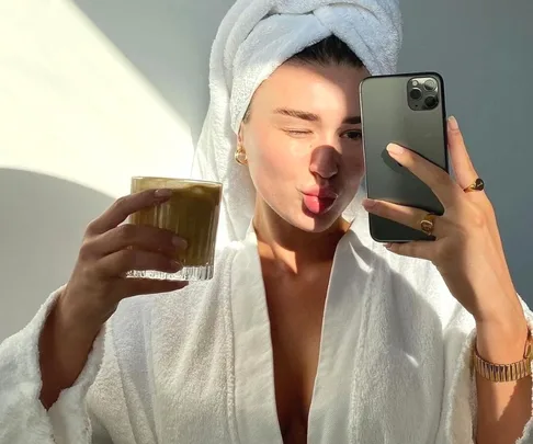 Person in a towel robe taking a selfie while holding a drink, with sunlight creating shadows on their face.