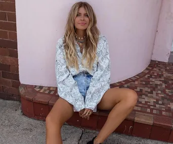 Woman with long hair in printed shirt and denim shorts sitting against a pink and brick wall background.