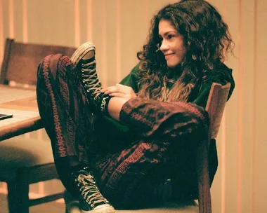 Rue from Euphoria sits relaxed with a smile, wearing patterned pants and sneakers, legs propped up on a chair.