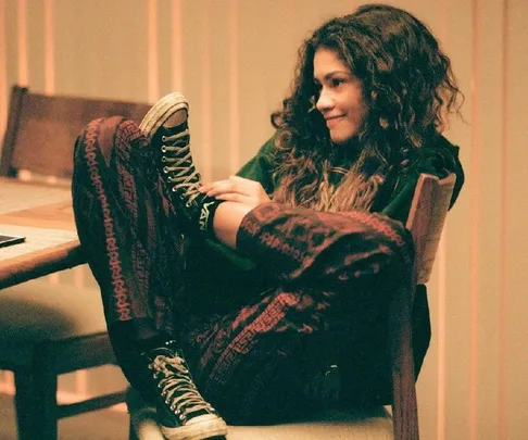 Rue from Euphoria sits relaxed with a smile, wearing patterned pants and sneakers, legs propped up on a chair.