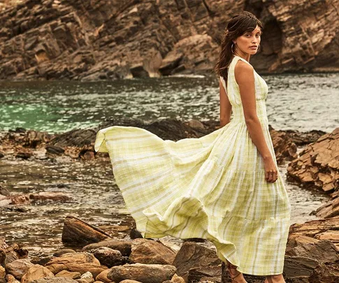 Woman in a flowing yellow dress stands on rocky shore, looking back, with ocean waves in the background.