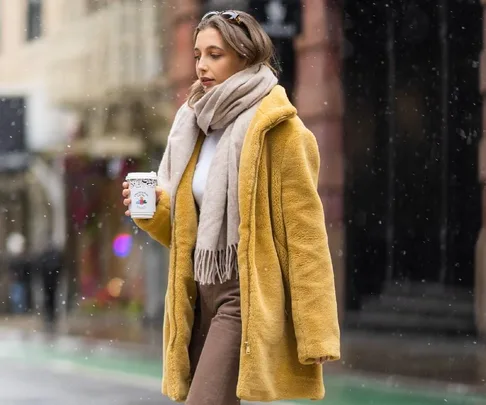 Woman in yellow coat and scarf walks in snowfall, holding a coffee cup, during New York Fashion Week street style.