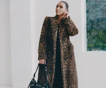 Person wearing a long leopard print coat, holding a black handbag, stands in a minimalist setting with a window.