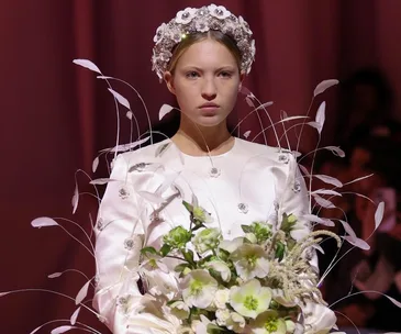 Model in white floral dress with decorative headpiece holding a bouquet on a runway.