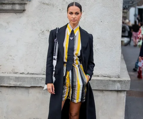 Street style at Milan Fashion Week Fall 2022; model in striped outfit with black coat and tie, standing against a wall.